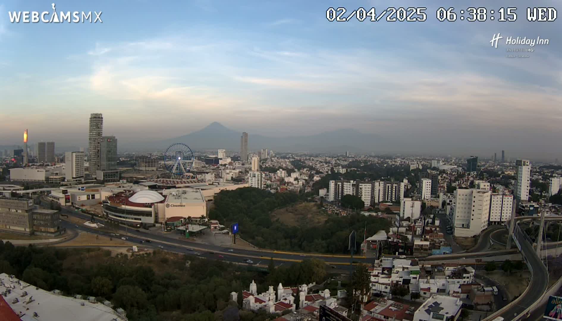 A hazy cityscape with a Ferris wheel and distant volcano is viewed from above under a mostly clear sky.