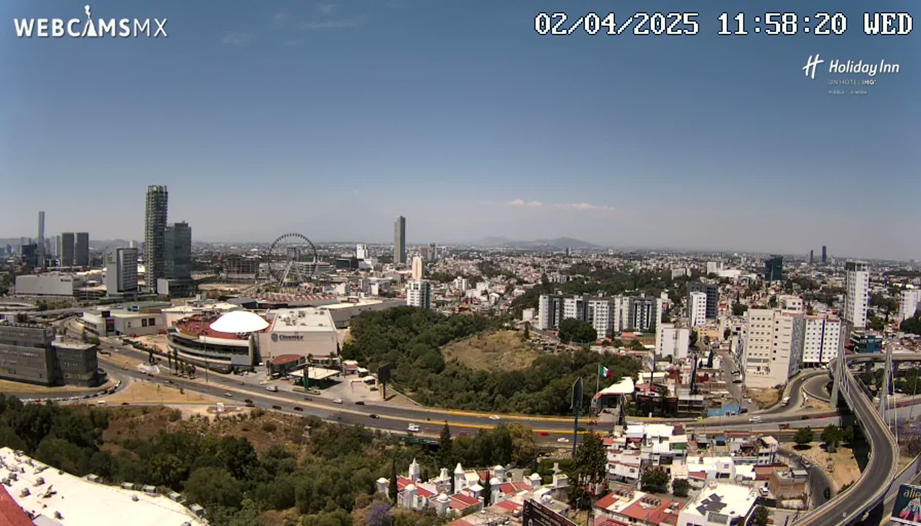 An aerial view of a city on a sunny day shows a large shopping mall, Ferris wheel, and highway interchange, surrounded by numerous buildings and green spaces.