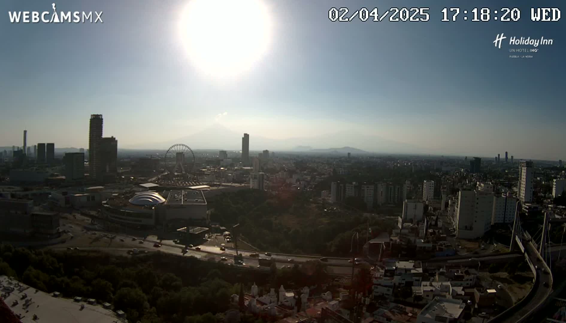 A sunny, hazy aerial view of a city featuring a Ferris wheel, several skyscrapers, and a highway curving through a residential area.