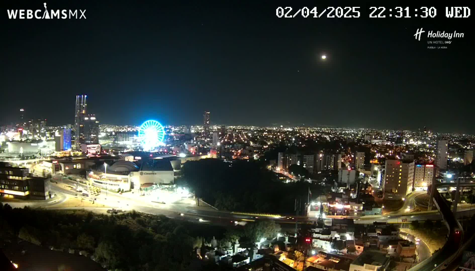 A nighttime, high-angle view of a city illuminated by lights, featuring a Ferris wheel and a partially visible moon in the clear sky.