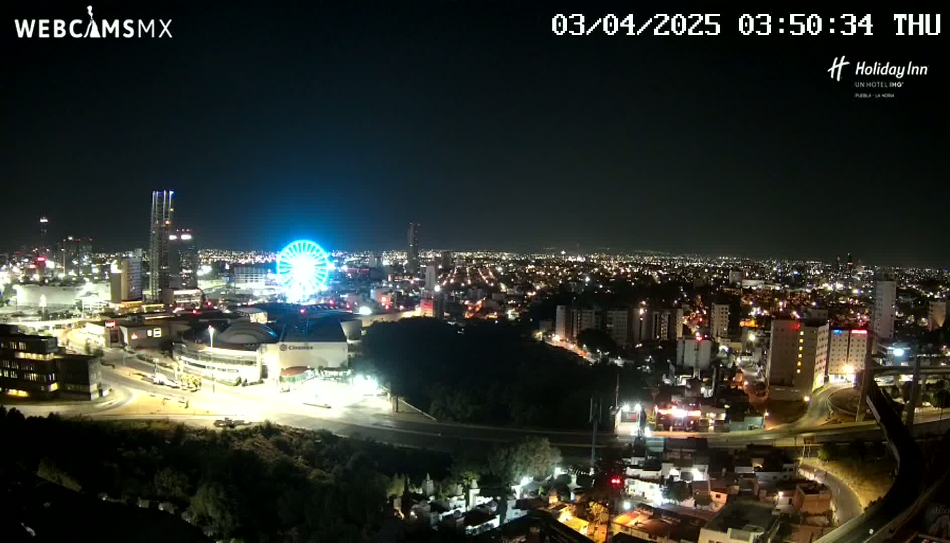A nighttime, aerial view of a city illuminated with lights, featuring a Ferris wheel and several buildings, under a clear sky.