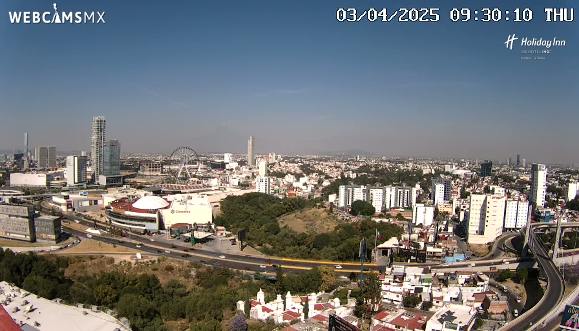 A sprawling city landscape under a clear blue sky, featuring a Ferris wheel, stadium, and several high-rise buildings.