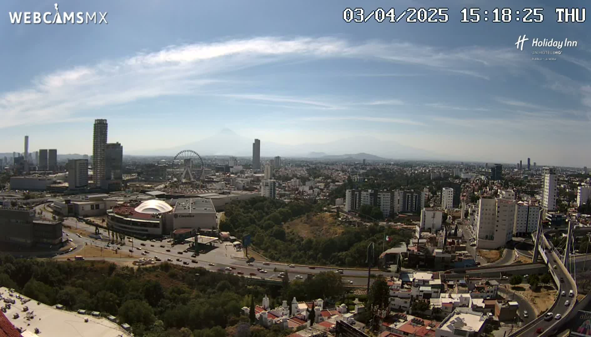 A sprawling city landscape under a mostly sunny sky with some scattered clouds is visible from a high vantage point, showcasing a large shopping mall, a Ferris wheel, and numerous high-rise buildings.