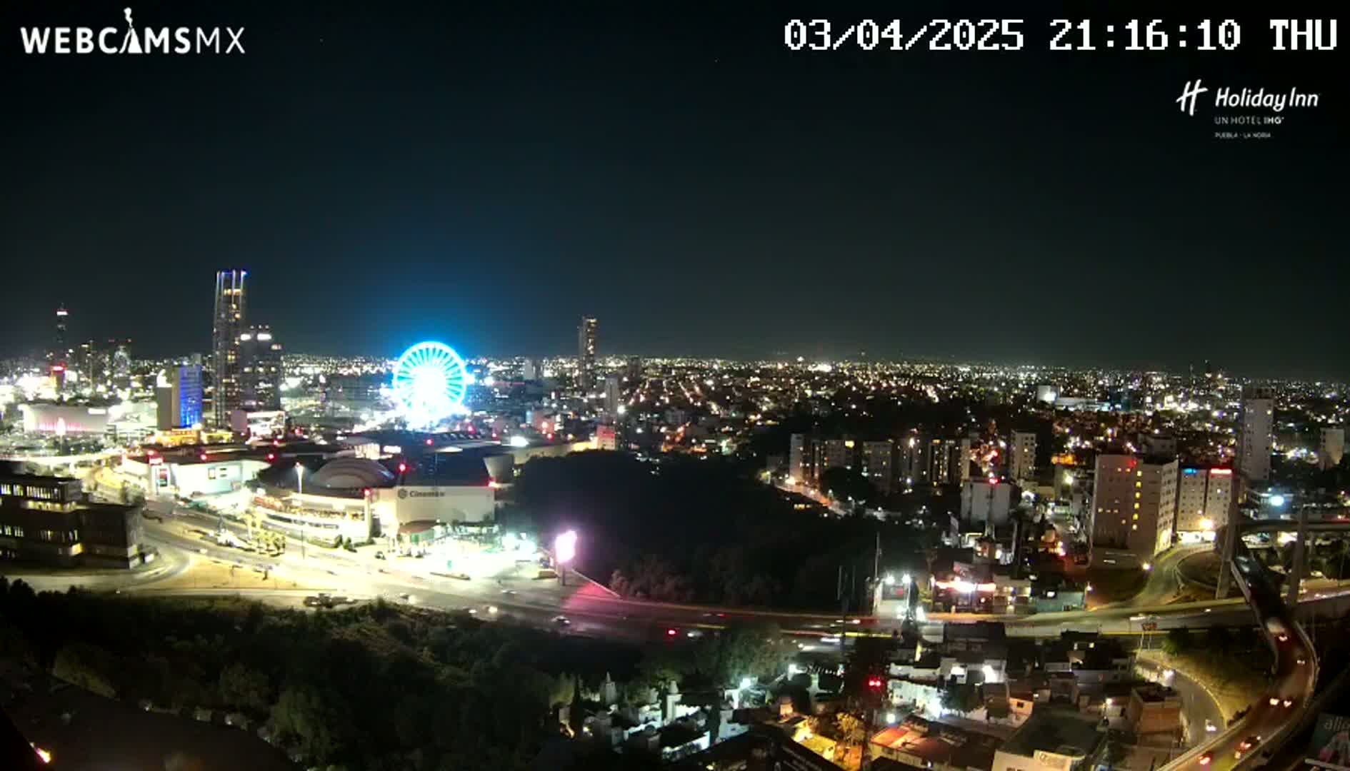 A nighttime aerial view of a city illuminated with lights, featuring a Ferris wheel and a large shopping complex, under a clear sky.