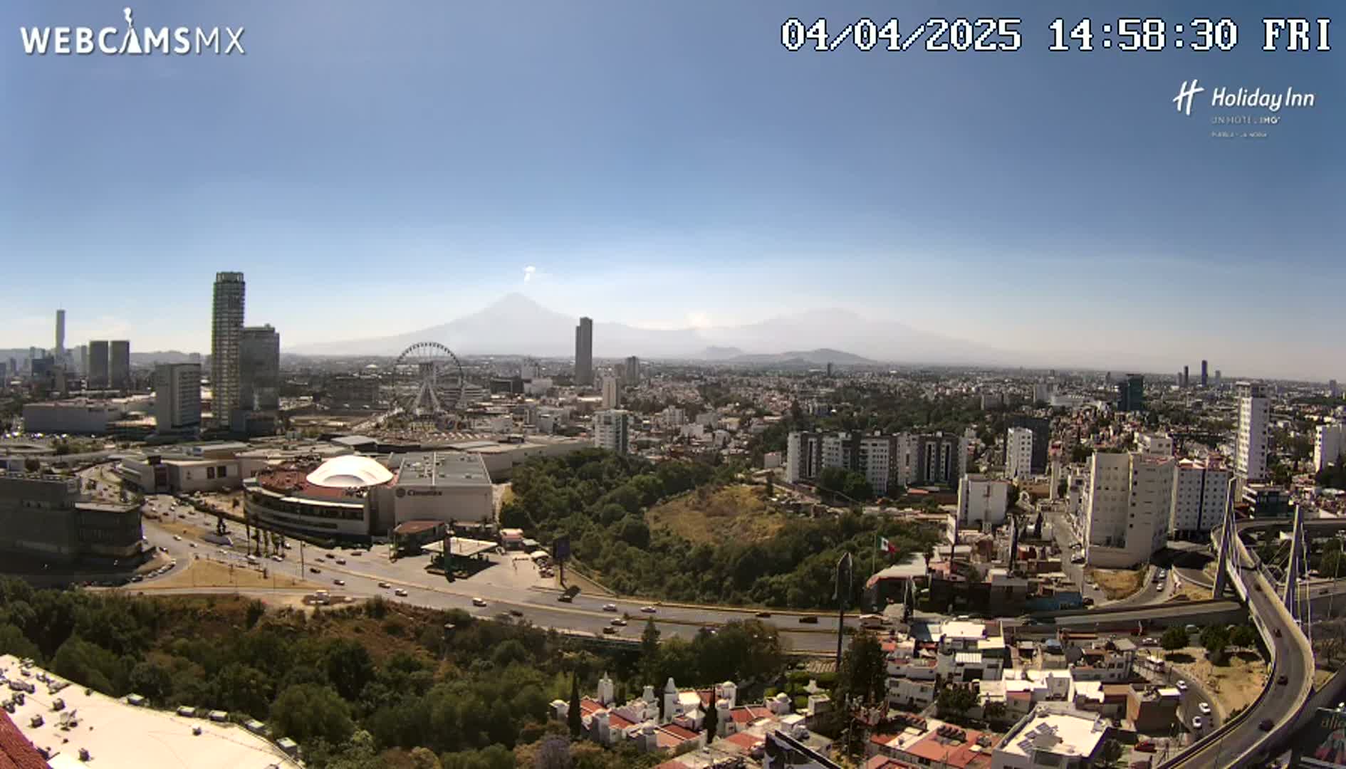 A high-angle, long shot of a sprawling city on a clear, sunny day, featuring a Ferris wheel, several skyscrapers, a large shopping mall, and a volcano visible in the distance.