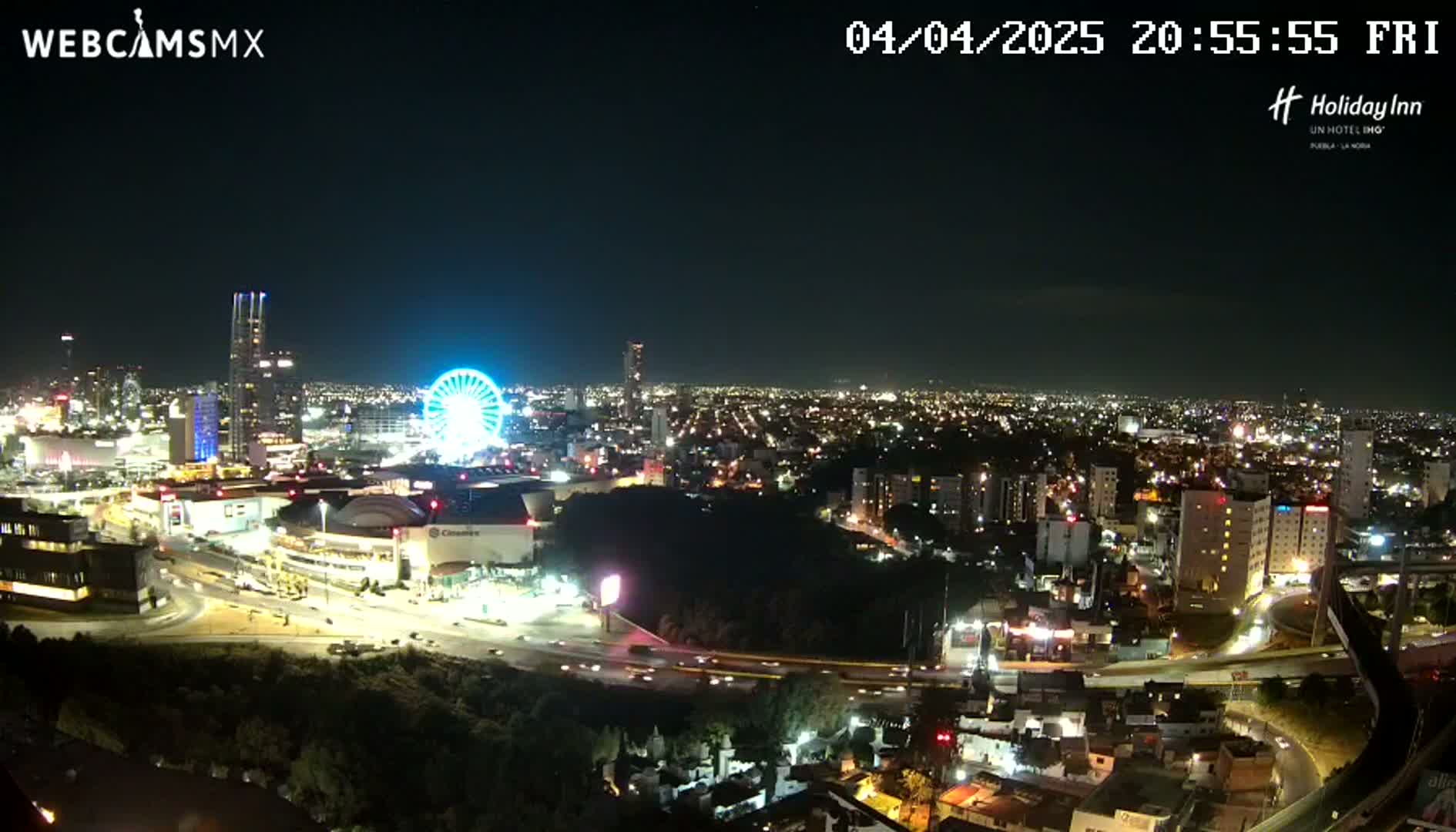 A nighttime aerial view of a city illuminated by streetlights, featuring a Ferris wheel and a large shopping mall, under a clear sky.