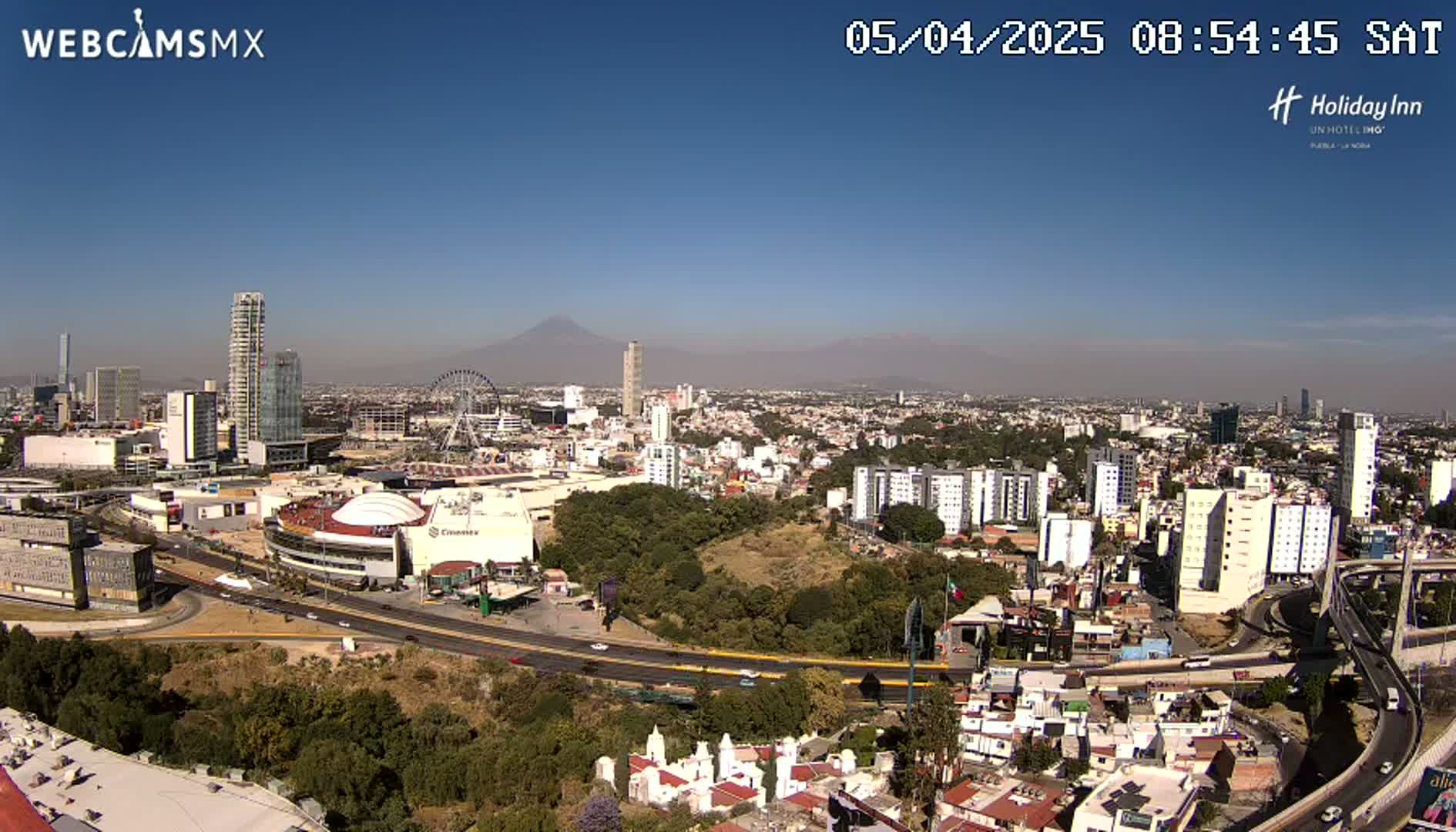 A sprawling city with numerous high-rise buildings, a Ferris wheel, and a large stadium is visible under a clear, sunny sky, with a volcano in the distance.