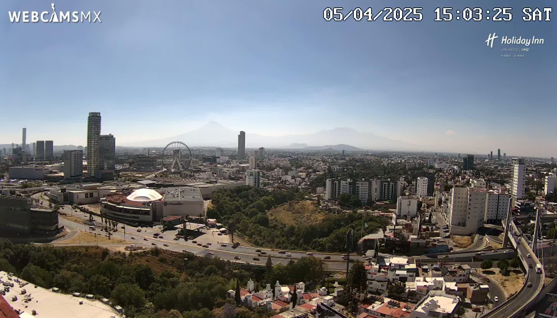 A sprawling city landscape under a clear, sunny sky, featuring a Ferris wheel, several high-rise buildings, and distant mountains visible on the horizon.