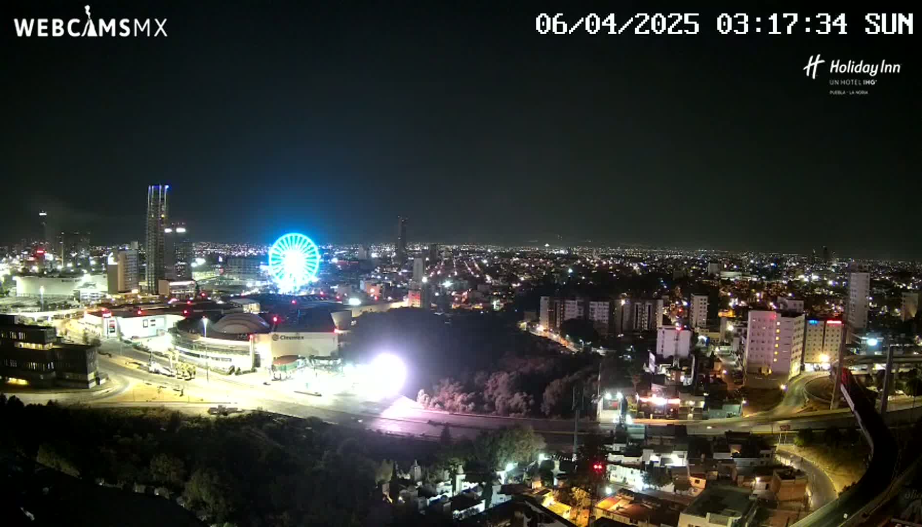 A nighttime, aerial view of a city with a Ferris wheel and brightly lit buildings, under a clear sky.