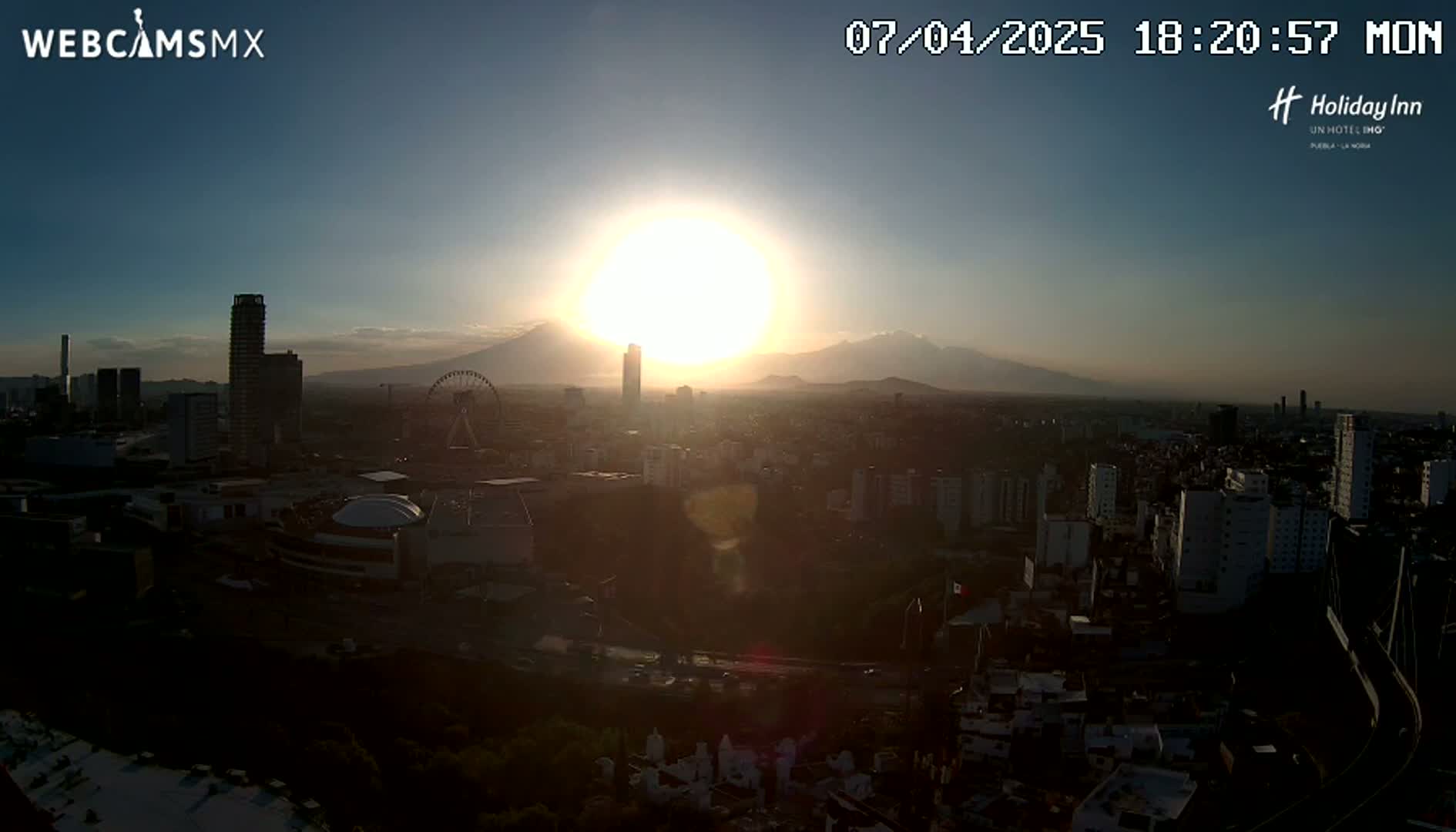 A bright sun sets over a city with a Ferris wheel and mountains in the distance.