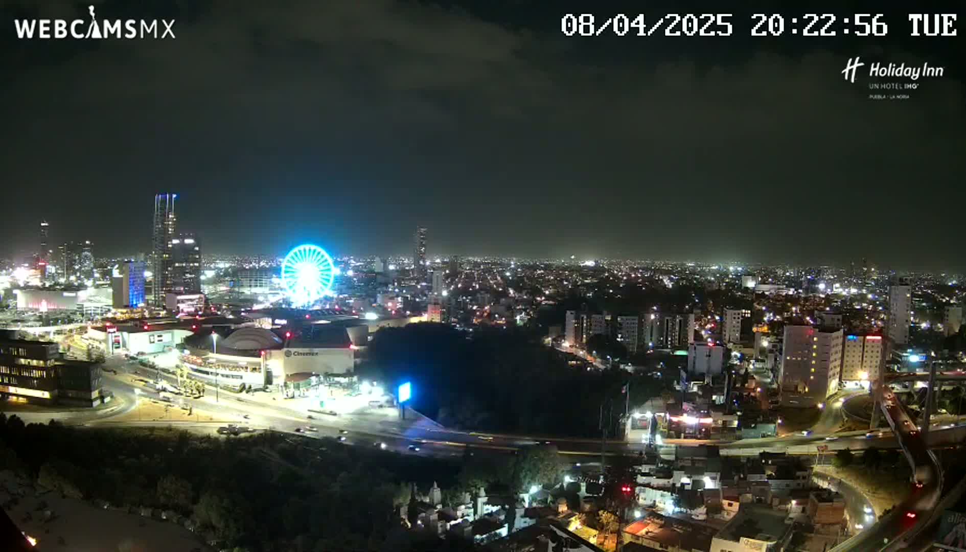 A nighttime cityscape with a Ferris wheel and many illuminated buildings under a partly cloudy sky.