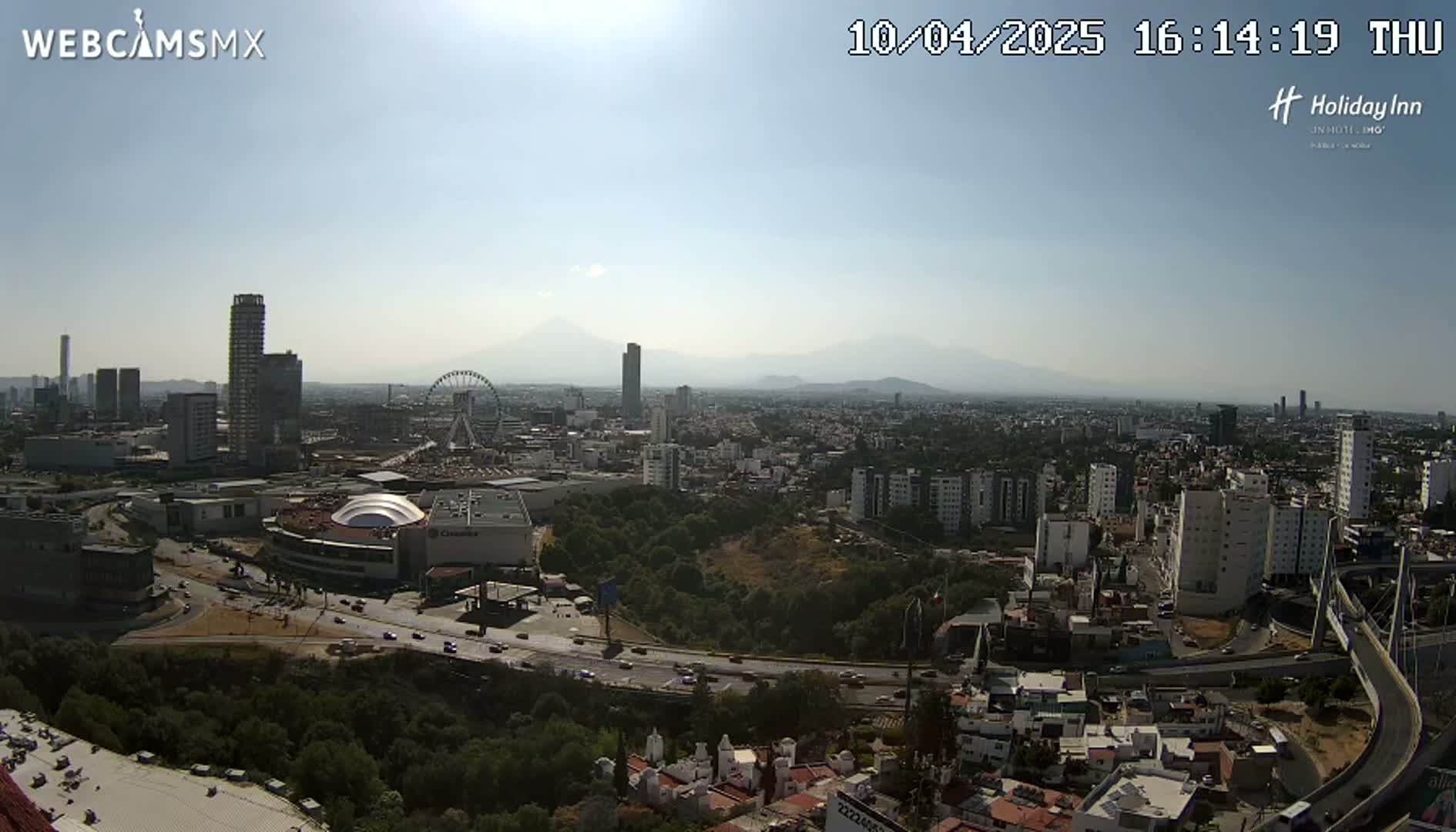 A sunny, hazy day reveals a sprawling cityscape with numerous buildings, a Ferris wheel, a large shopping mall, and mountains in the distance.