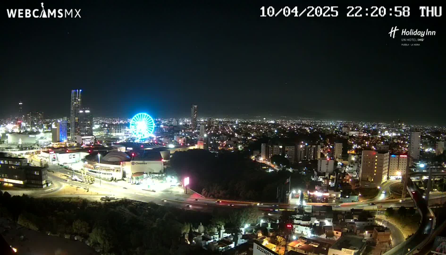 A nighttime, aerial view of a city illuminated with lights, featuring a Ferris wheel and several buildings, under a clear sky.