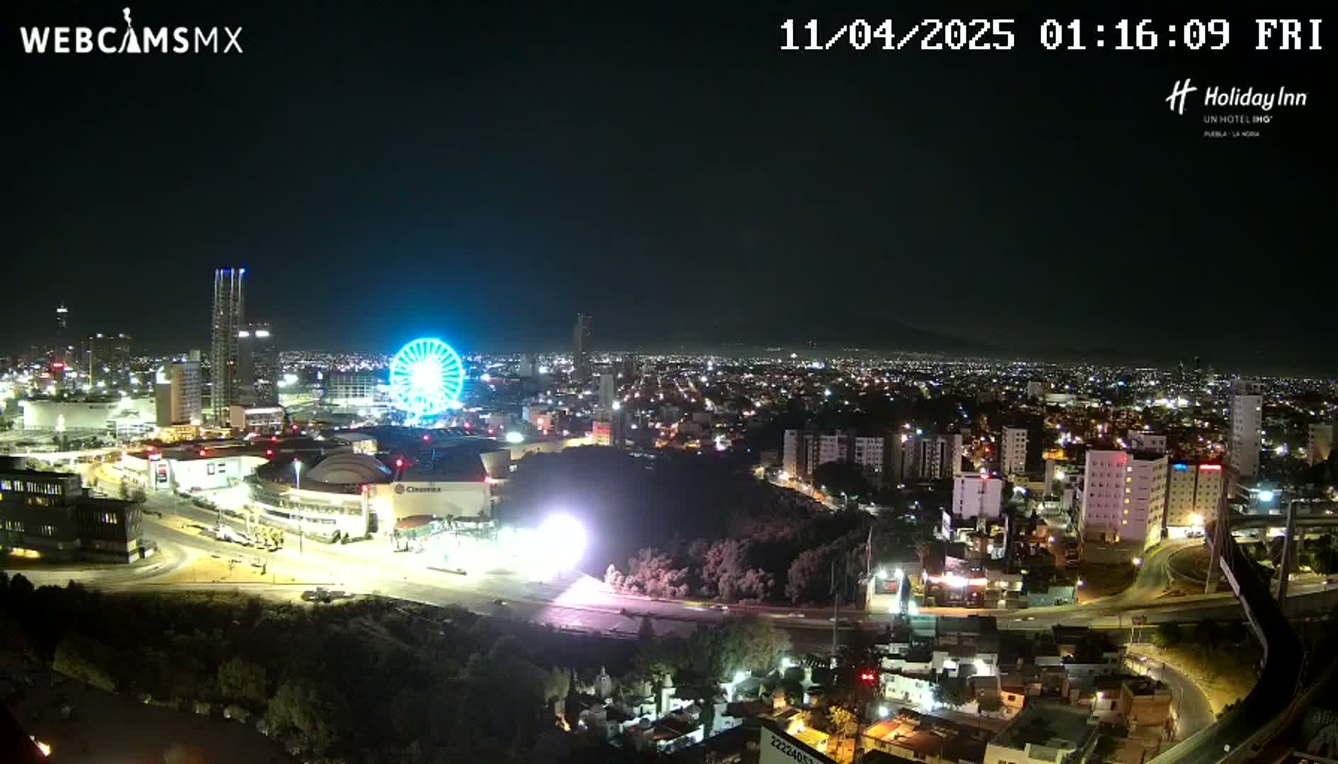 A nighttime, aerial view of a city illuminated by lights, featuring a Ferris wheel and a large building complex.