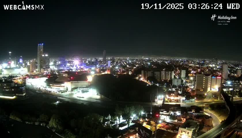 A wide aerial view shows a brightly lit city at night under a clear sky, featuring numerous buildings, roads, and a prominent Ferris wheel in the distance.