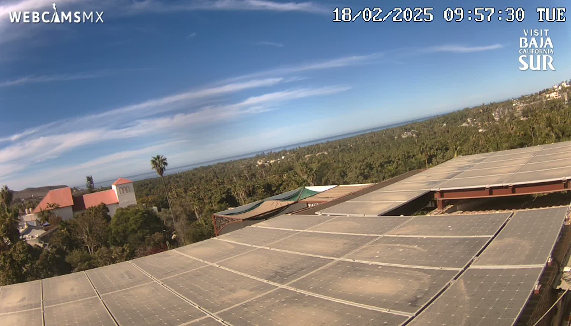 A large solar panel array on a building overlooks a coastal town and ocean under a mostly clear blue sky.