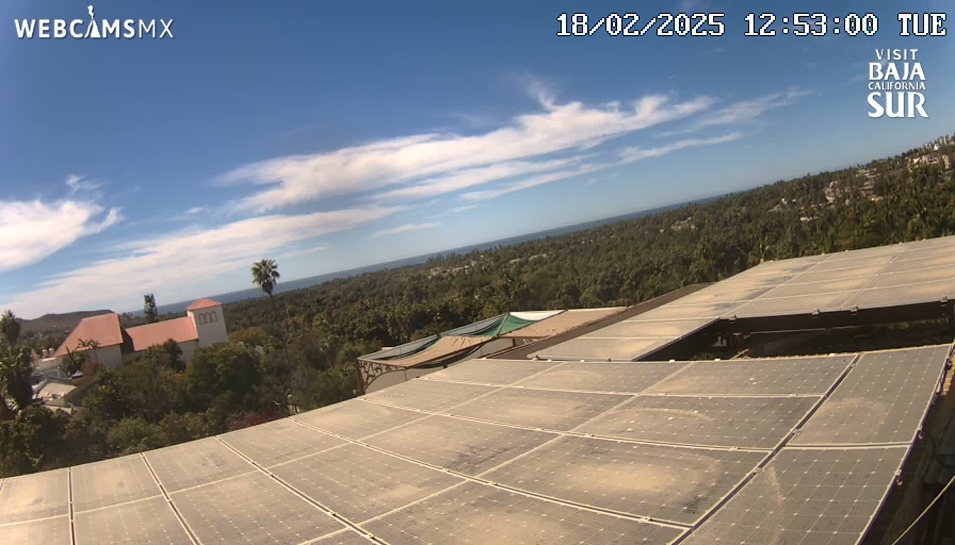 A mostly sunny day overlooks a sprawling landscape of trees, a building with a red roof, and a large expanse of solar panels.