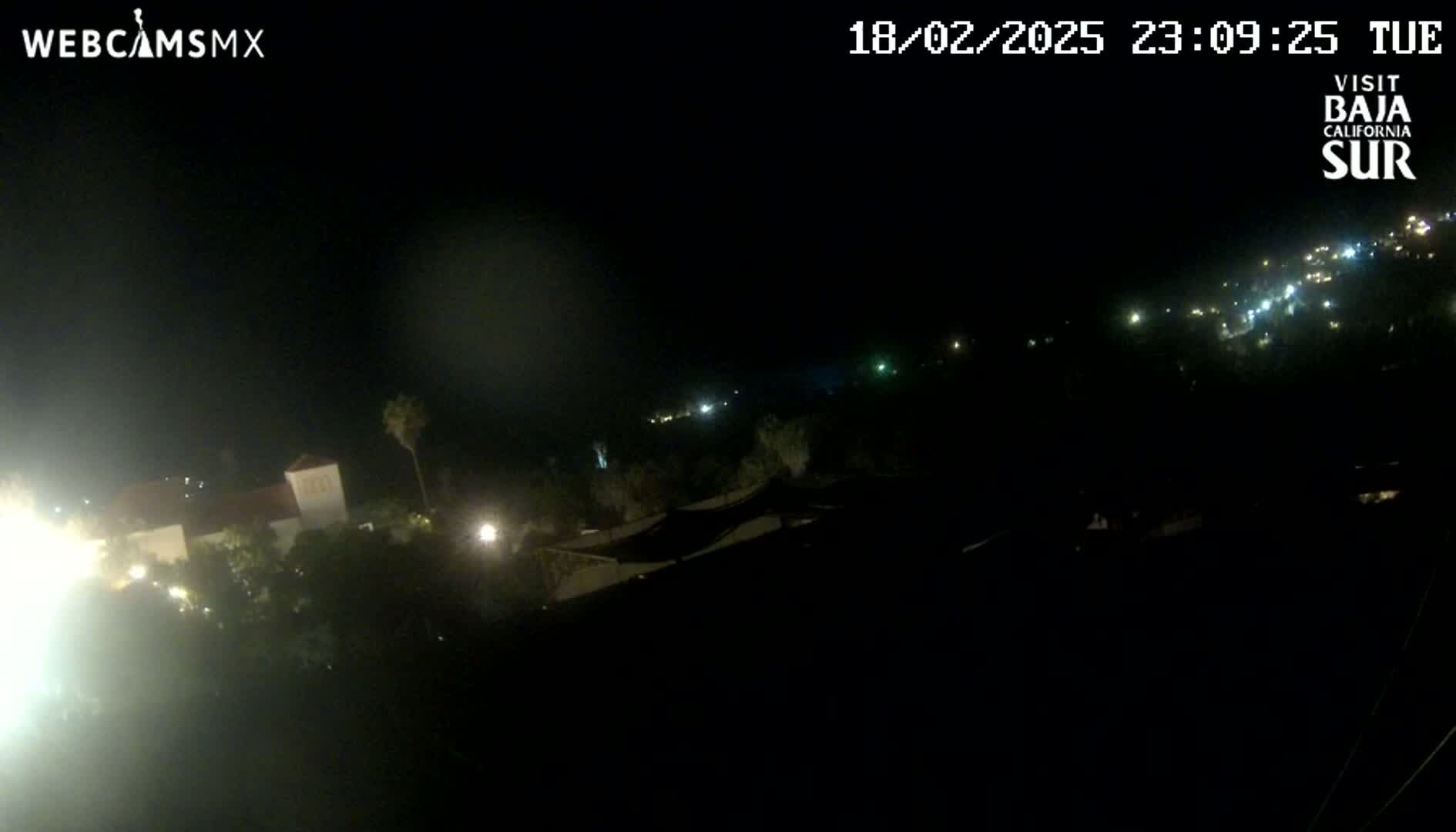 A nighttime, high-angle view shows a town with scattered lights, some buildings, and vegetation under a dark sky.