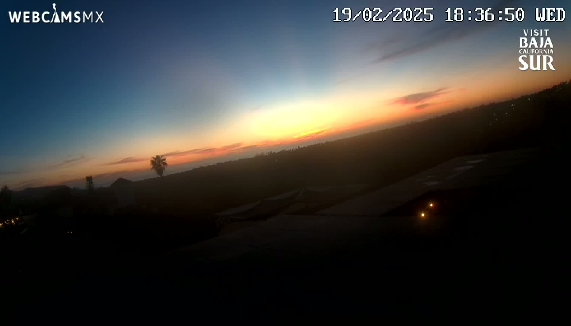 A sunset over a dark, hilly landscape with a few lights visible in the foreground.