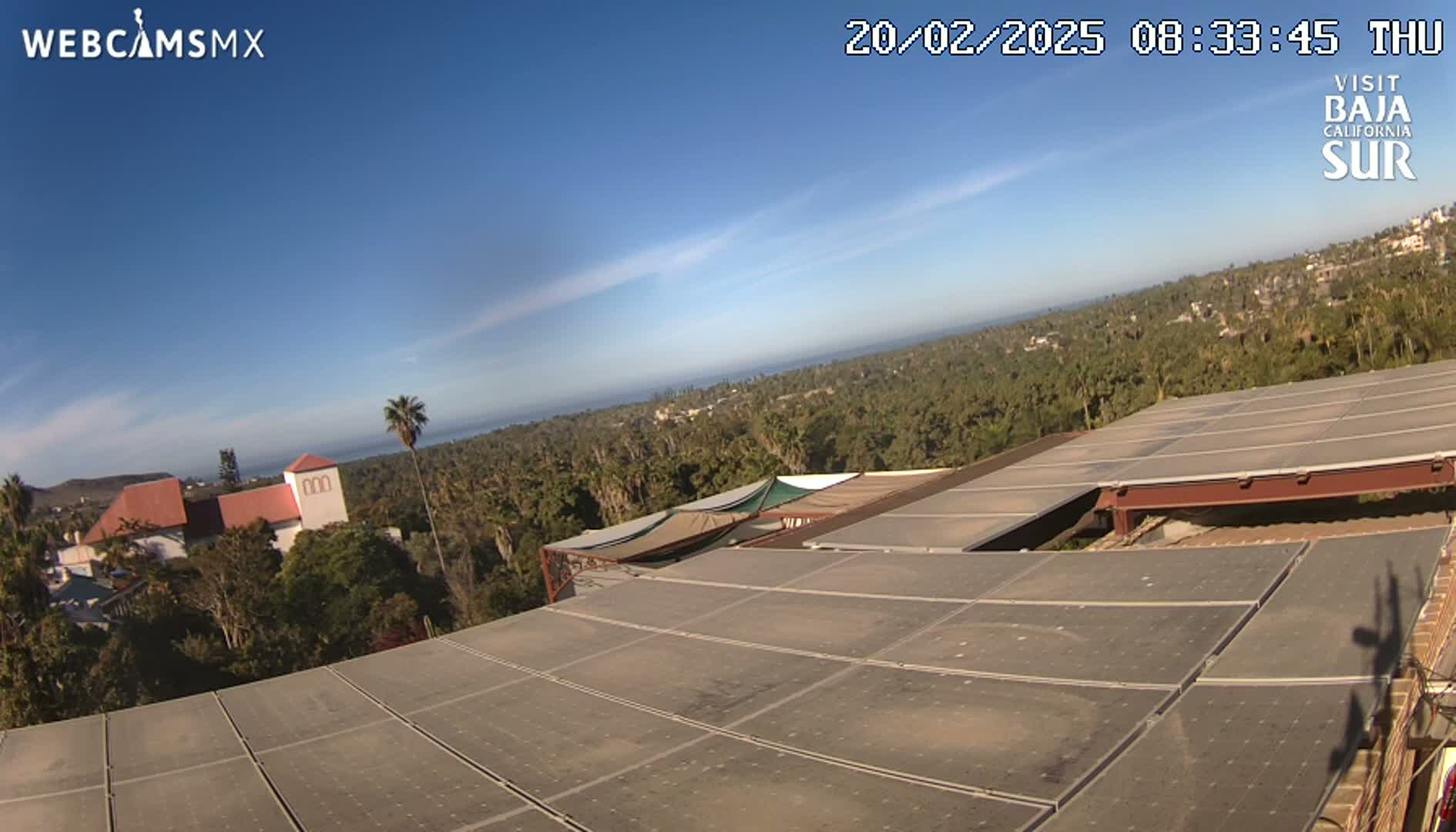A large solar panel array sits atop a building overlooking a coastal town, ocean, and clear blue sky.