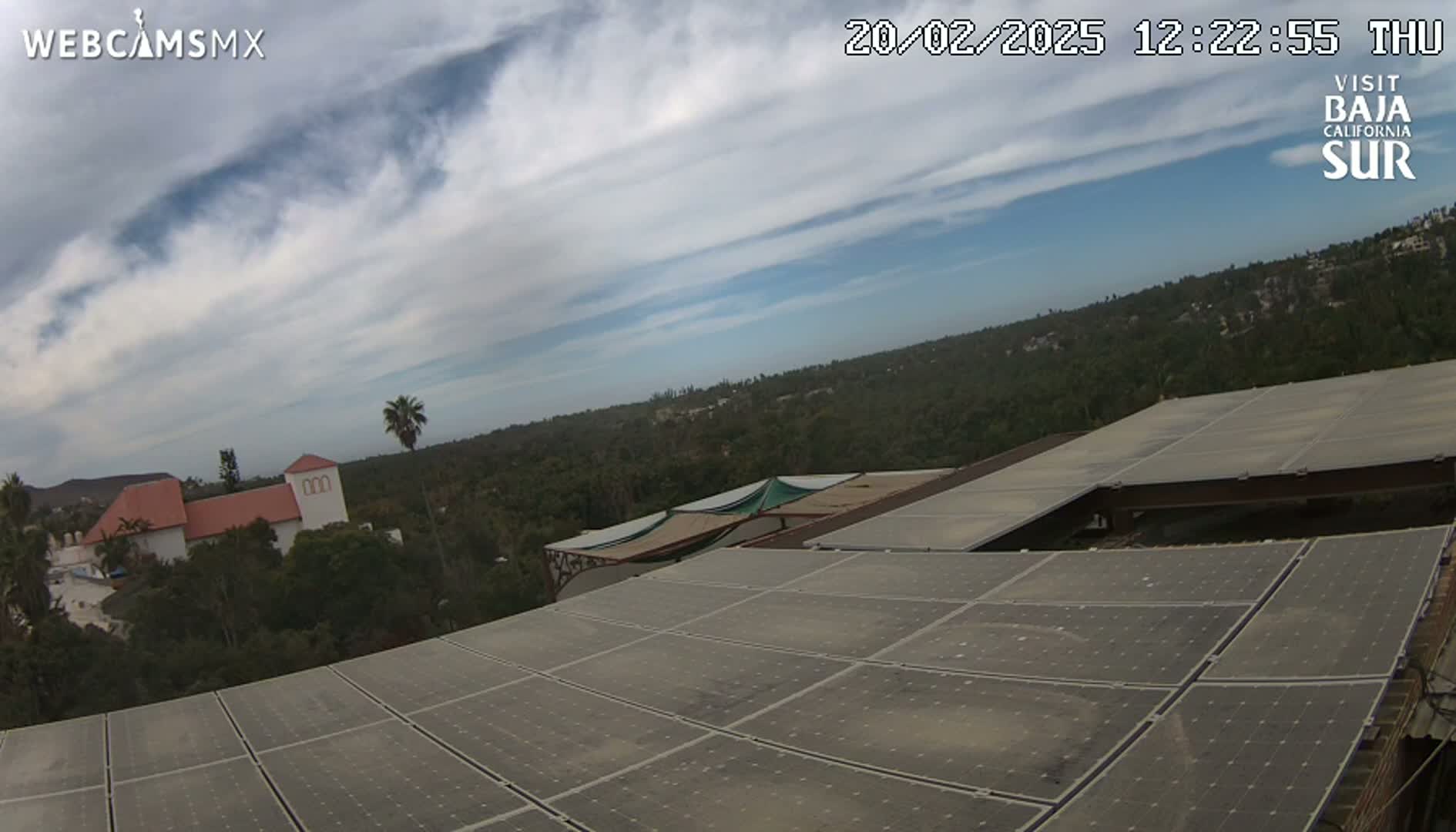 A partly cloudy sky overlooks a large solar panel array on a building, with a distant view of buildings and trees.