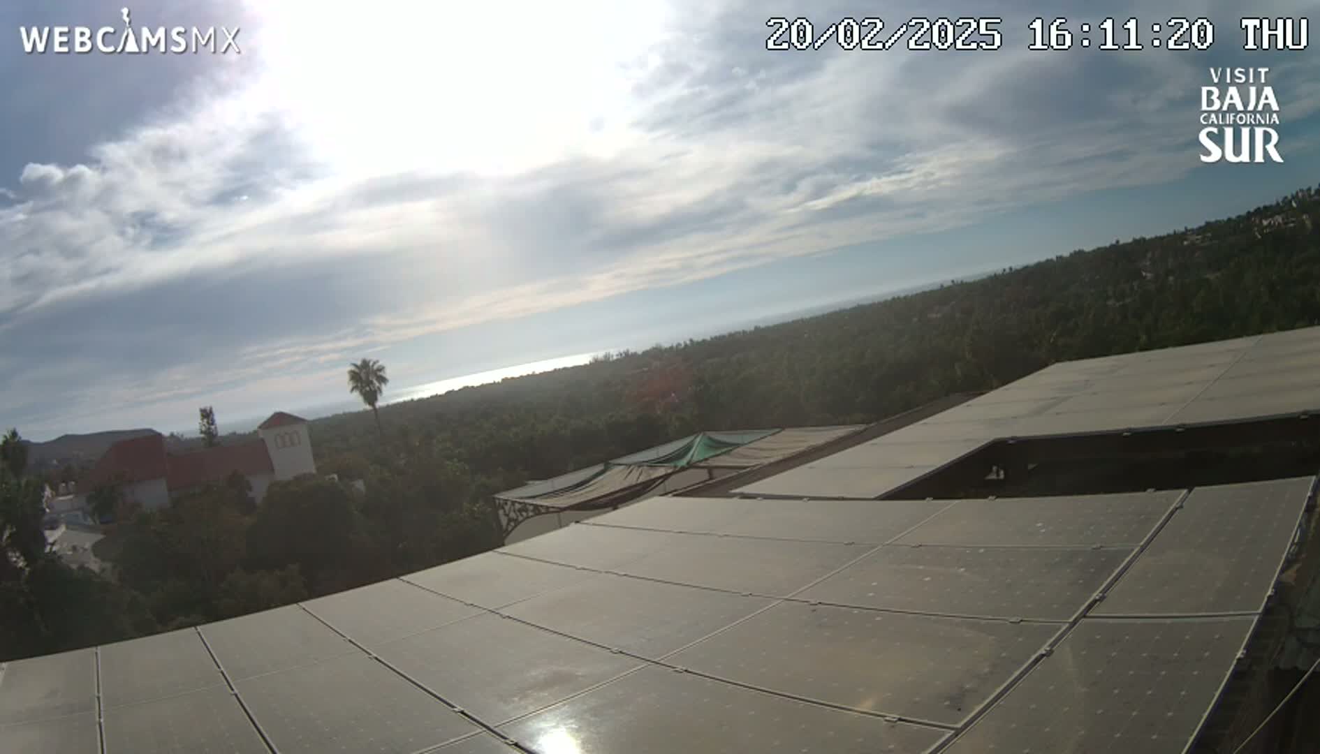 A partly cloudy sky overlooks a coastal landscape of trees and buildings, featuring a large solar panel array in the foreground.