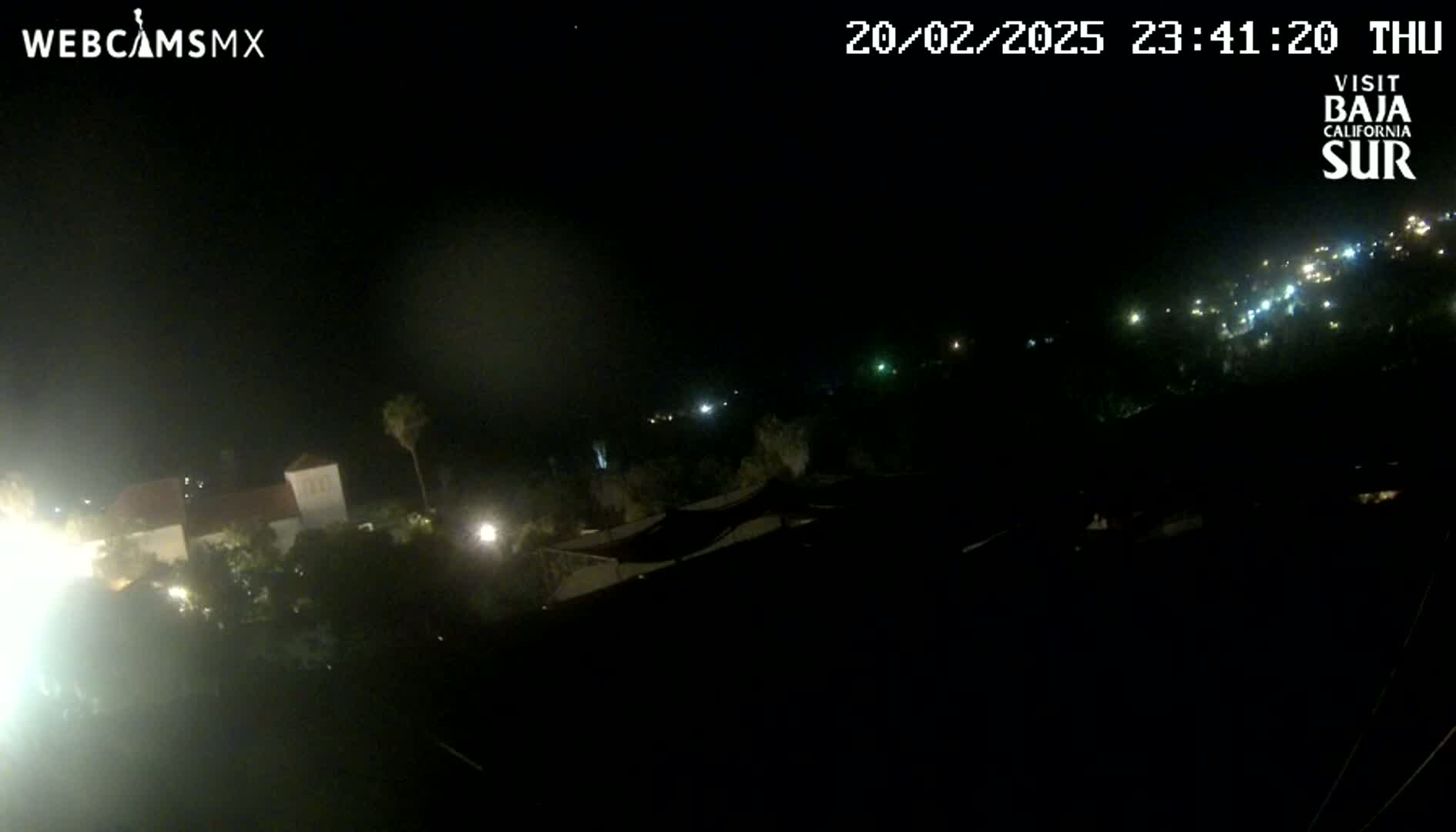 A nighttime, high-angle view shows a dark landscape with sparsely lit buildings and vegetation, under a clear, dark sky.