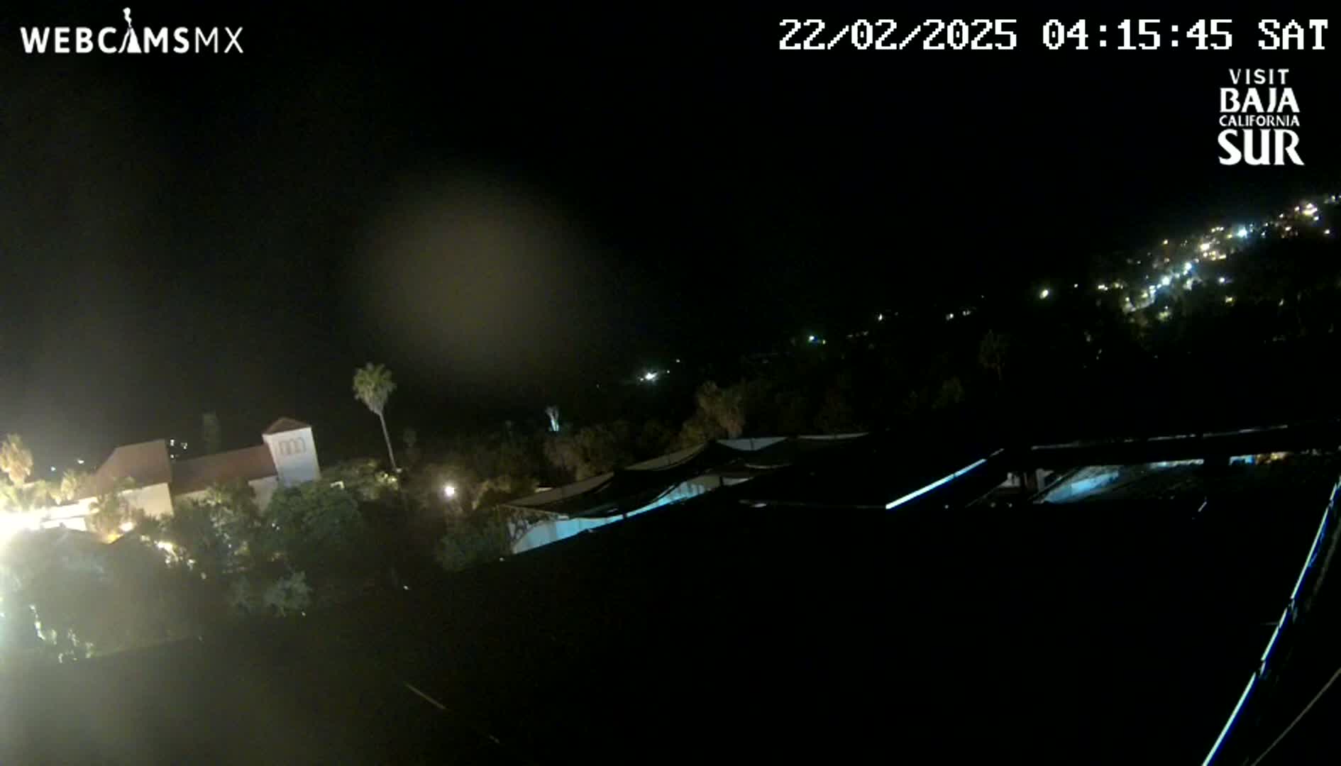 A nighttime, high-angle view shows a building complex and surrounding vegetation, with a distant town illuminated in the background under a clear, dark sky.