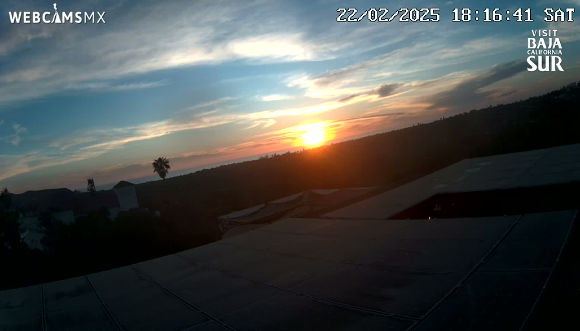 A sunset over a silhouetted landscape is viewed from a rooftop, with a mostly clear sky.