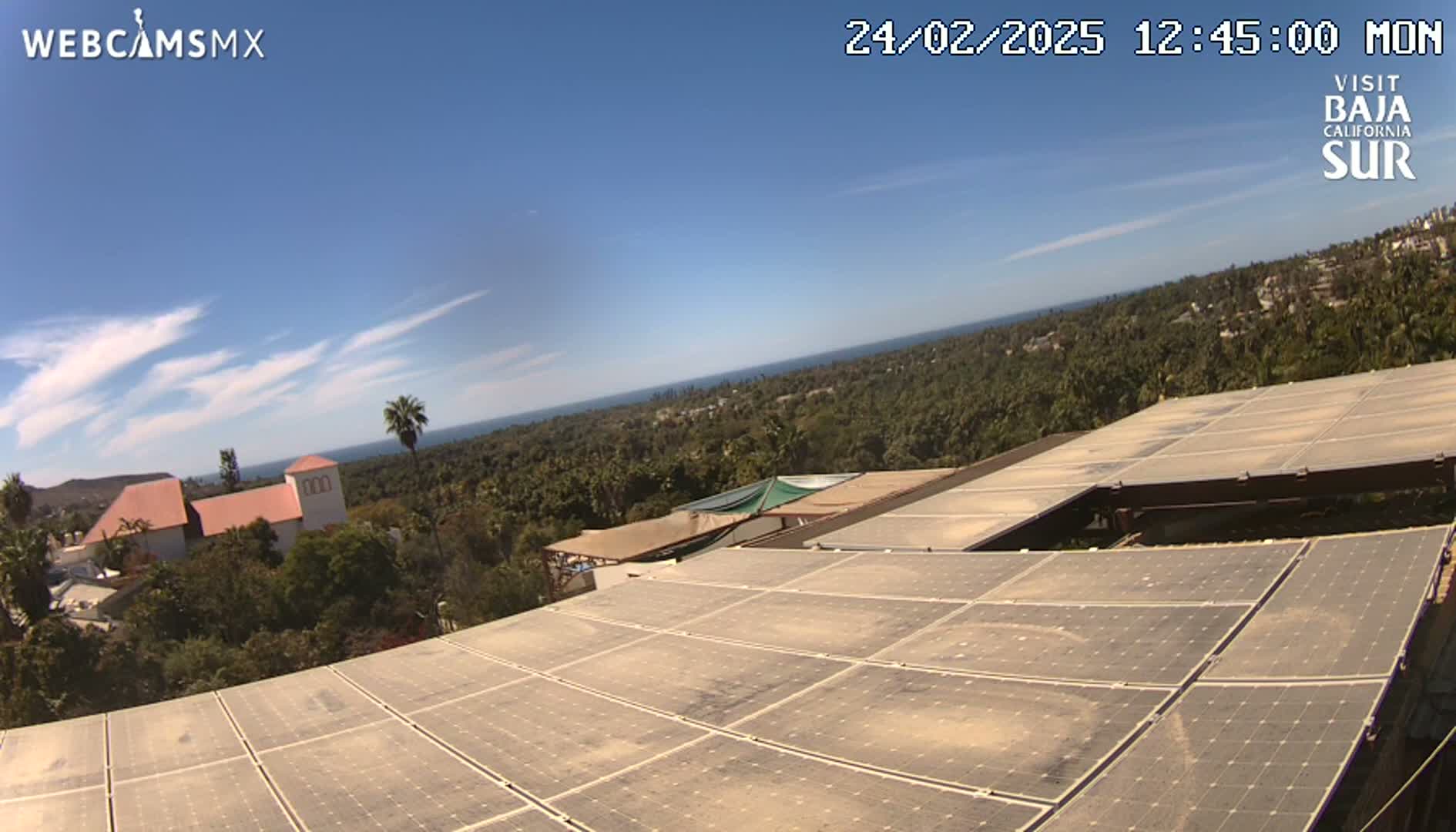 A large array of solar panels on a rooftop overlooks a coastal landscape under a clear blue sky with a few wispy clouds.