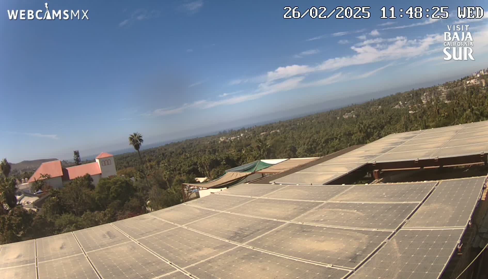 A mostly sunny day reveals a view of a dusty solar panel array on a rooftop overlooking a coastal town nestled among lush greenery and a distant ocean.