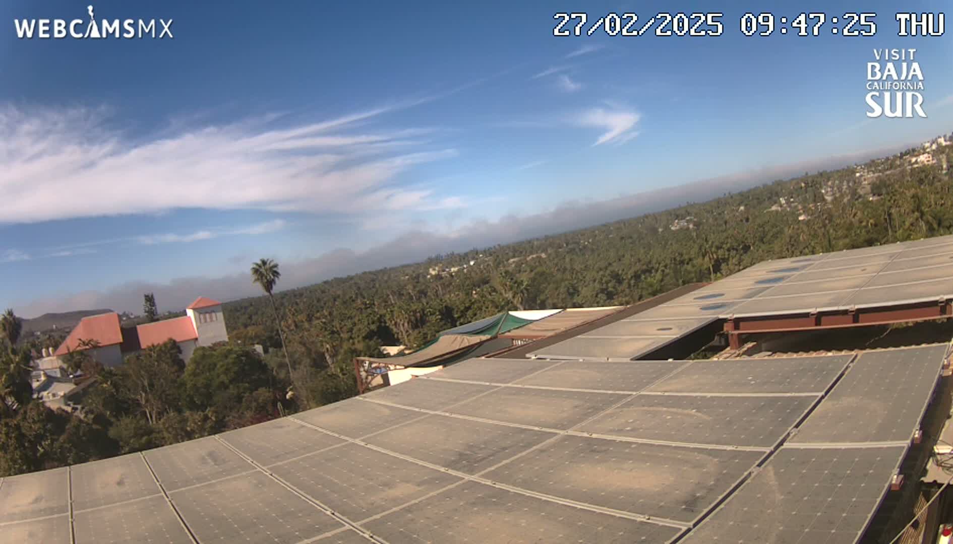 A mostly clear, sunny day reveals a rooftop covered in solar panels overlooking a town nestled amongst lush greenery, with hazy mountains visible in the distance.