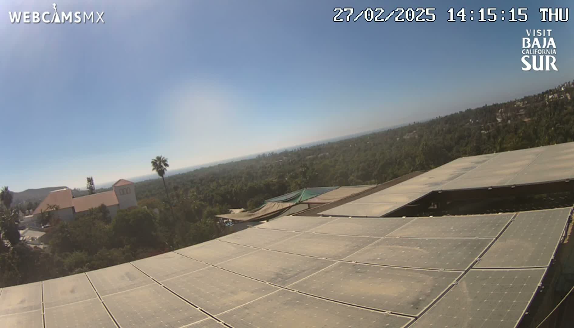 A large array of solar panels sits atop a building overlooking a sunny, tree-covered landscape that extends to a distant ocean.