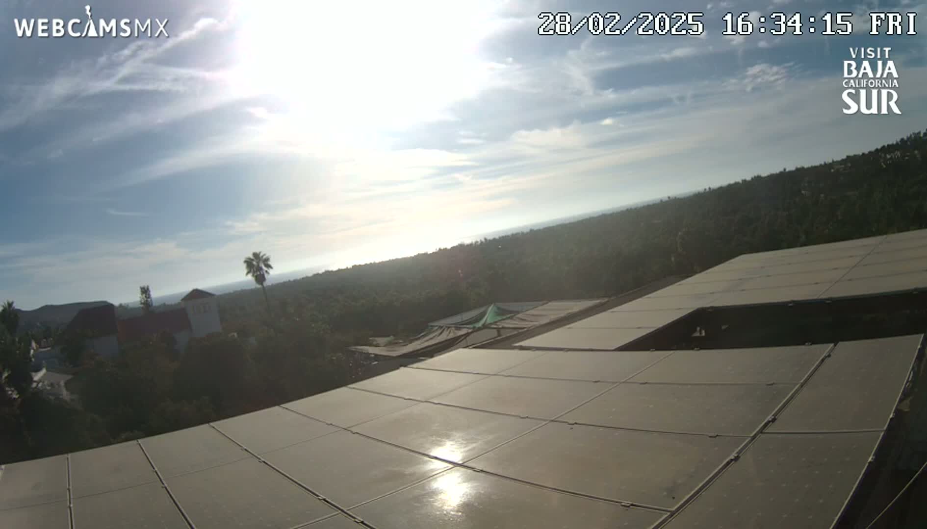 A mostly sunny day overlooks a hilly landscape of green vegetation and buildings, viewed from a vantage point on a large, flat, solar panel array.