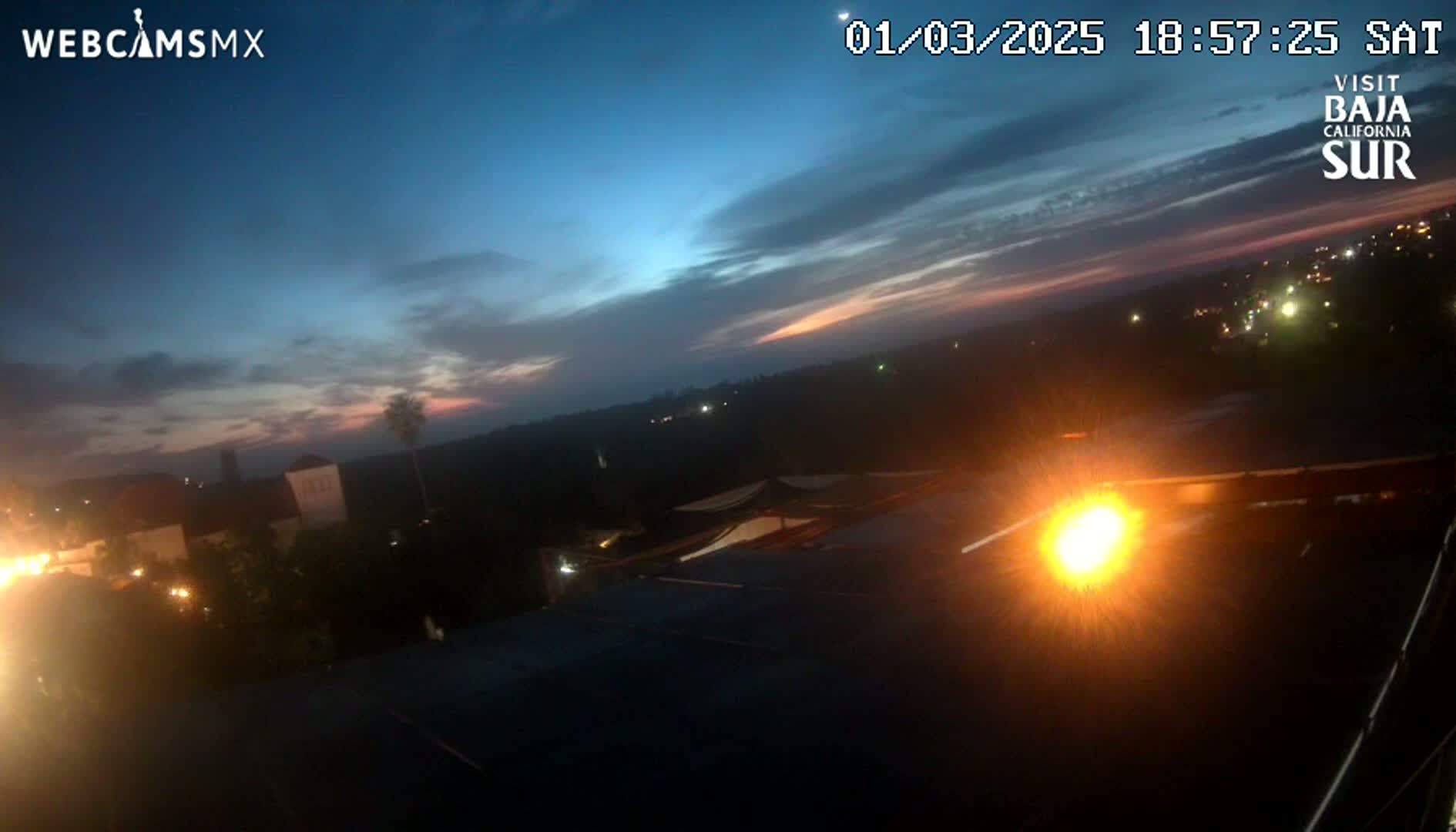 A twilight scene shows a partly cloudy sky with hints of pink and orange near the horizon, over a dark landscape with some lights visible in the distance, and bright headlights are visible in the foreground.
