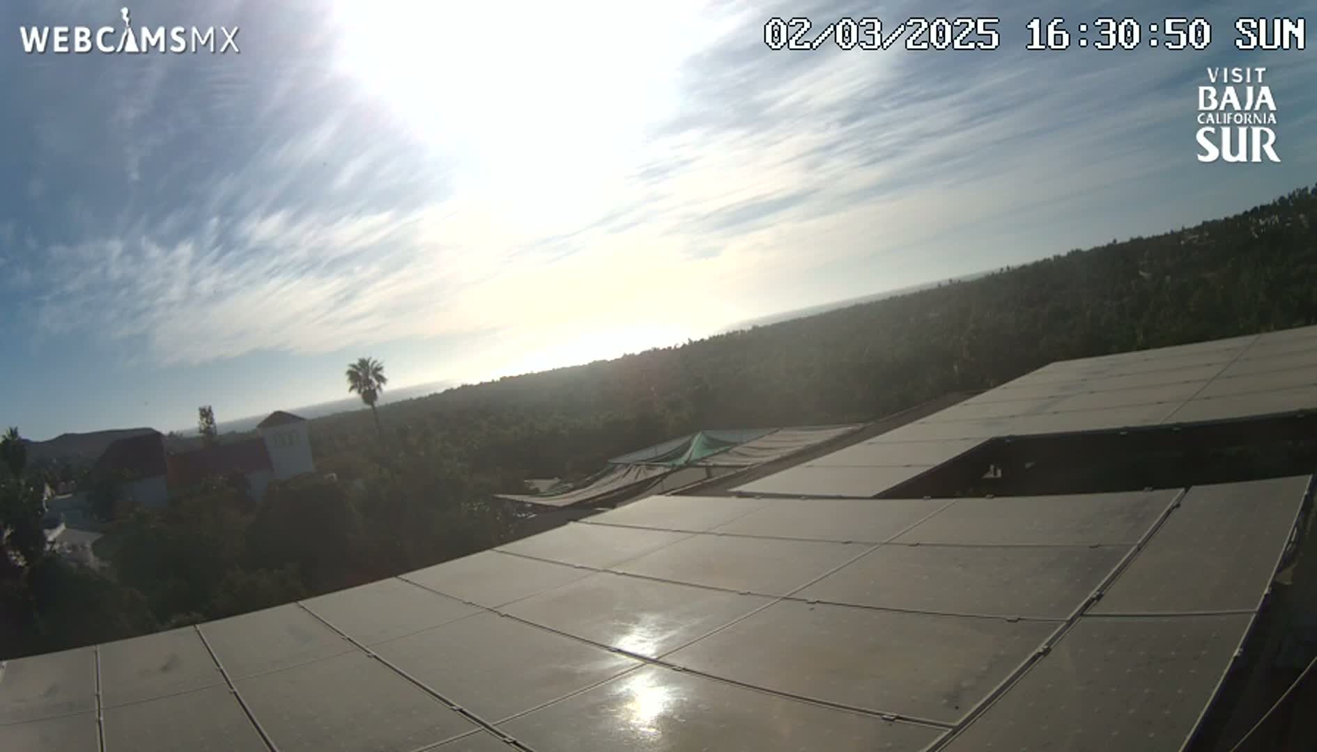 A mostly sunny day with light clouds over a hilly landscape, viewed from a rooftop covered in solar panels.