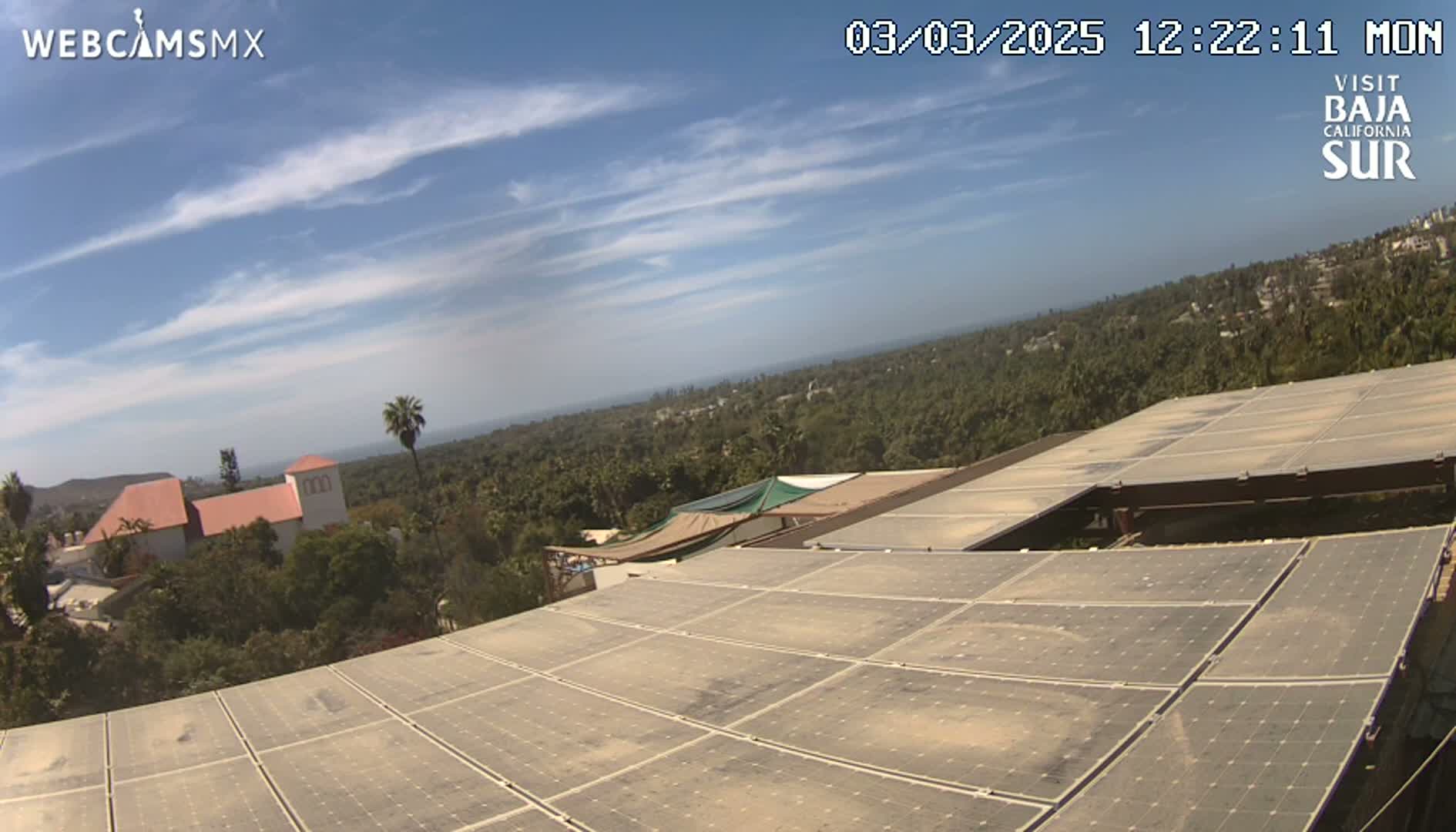 A mostly sunny day with a few thin clouds over a landscape of buildings and trees, viewed from a rooftop covered in solar panels.