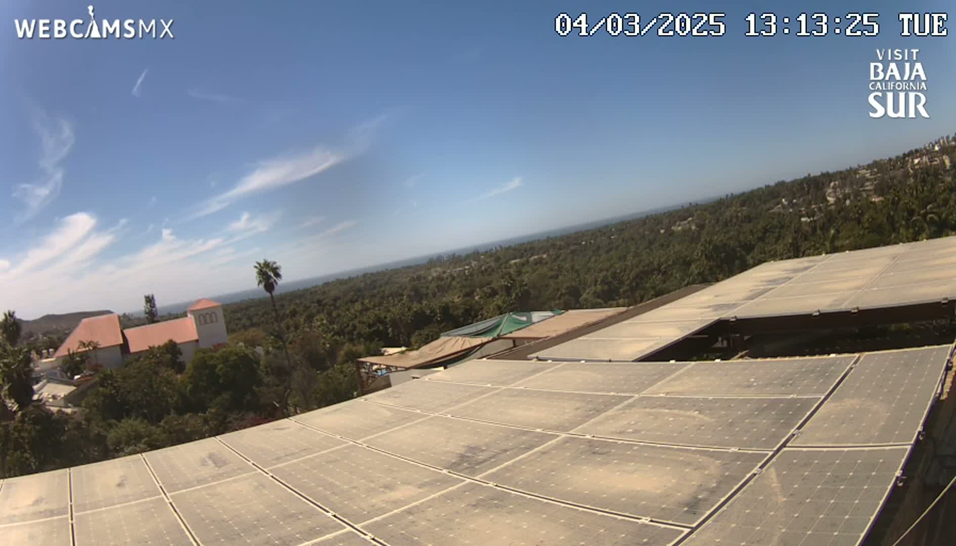 A large solar panel array sits atop a building overlooking a sunny, ocean-view landscape with lush green vegetation and distant buildings.
