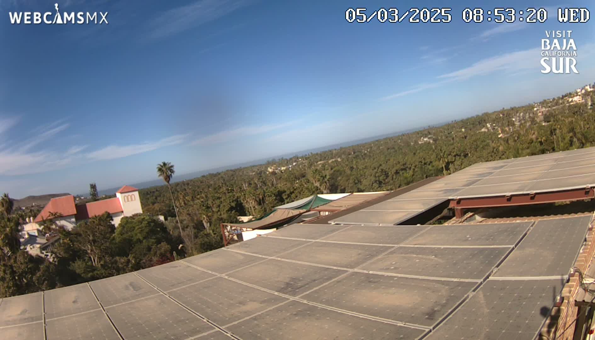 A large solar panel array on a building overlooks a clear, sunny day with a view of ocean and distant buildings surrounded by lush vegetation.