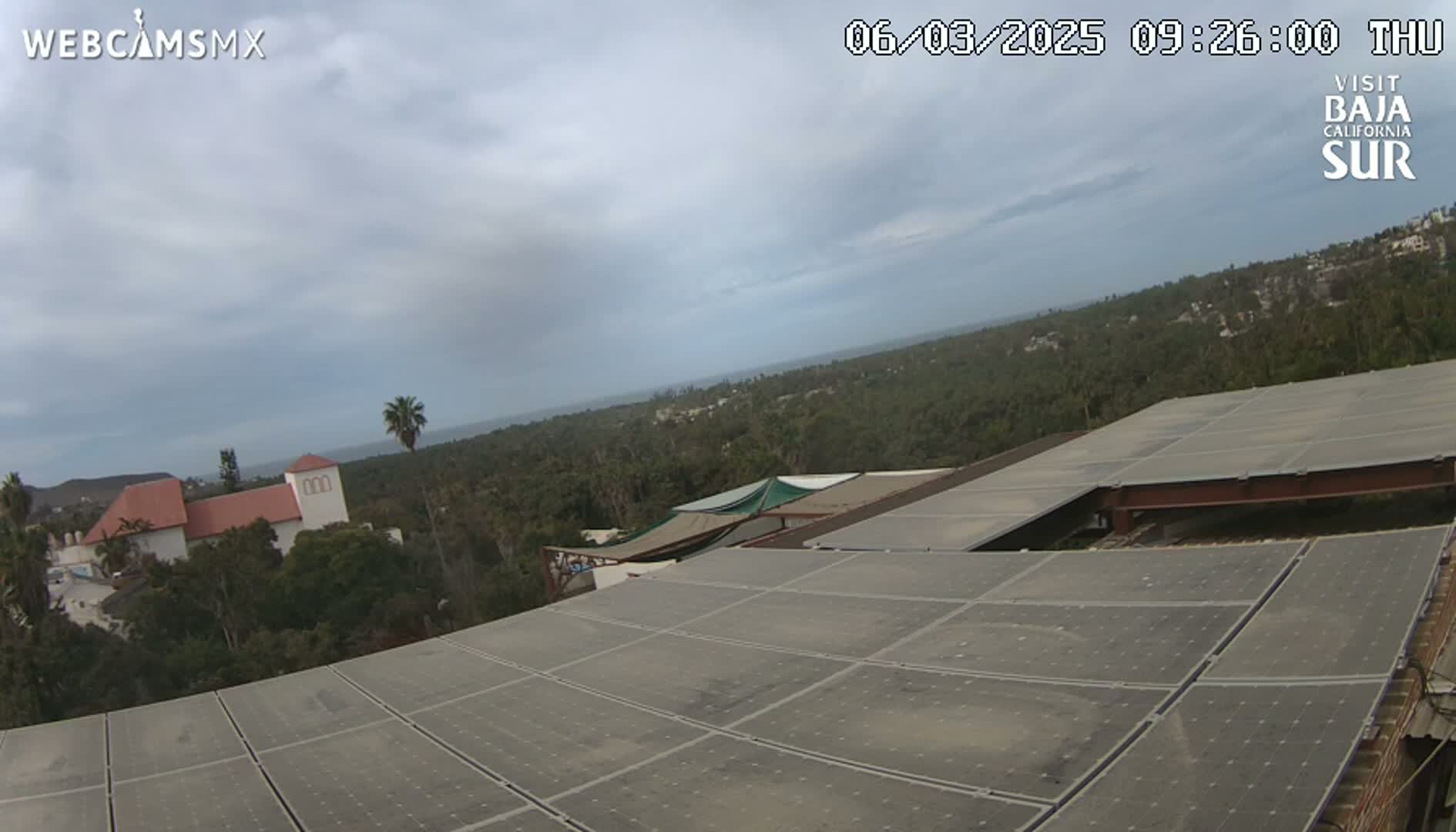 An overcast sky overlooks a landscape of lush green vegetation, a building with a red tile roof, and a large expanse of solar panels.