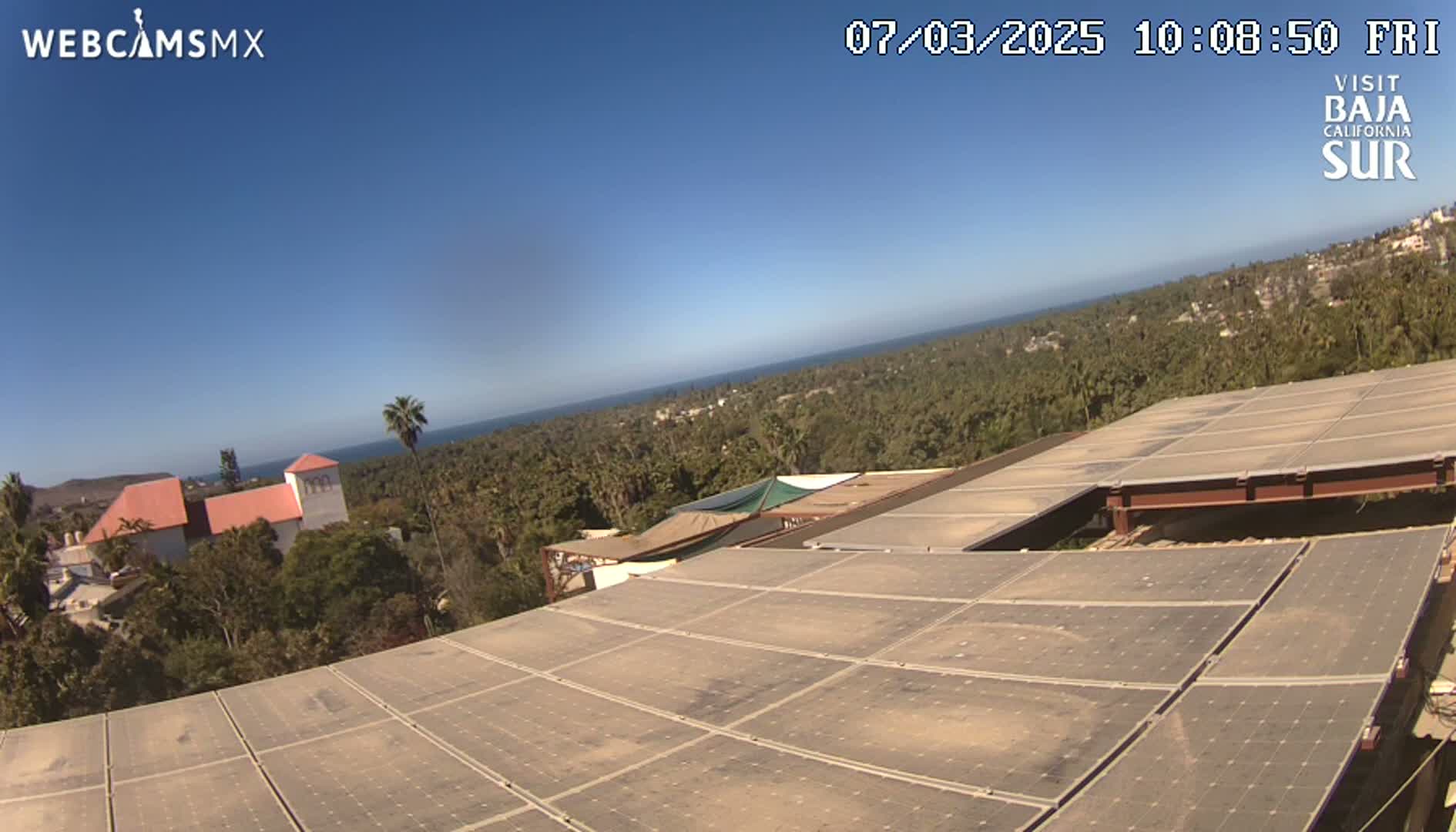 A large array of solar panels sits atop a building overlooking a coastal town and ocean under a clear blue sky.