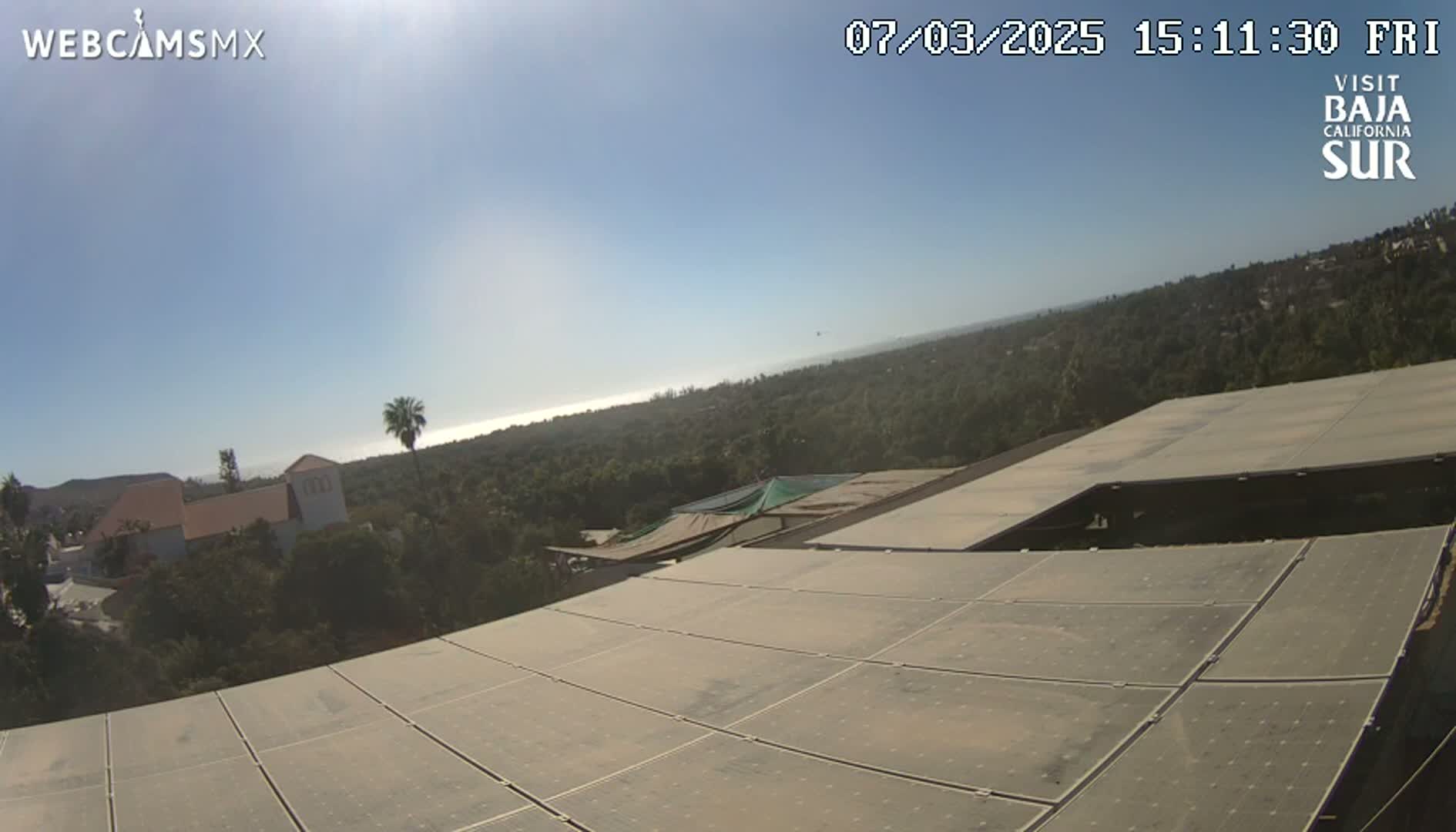 A large solar panel array on a building overlooks a tree-covered landscape and a distant ocean under a clear, sunny sky.