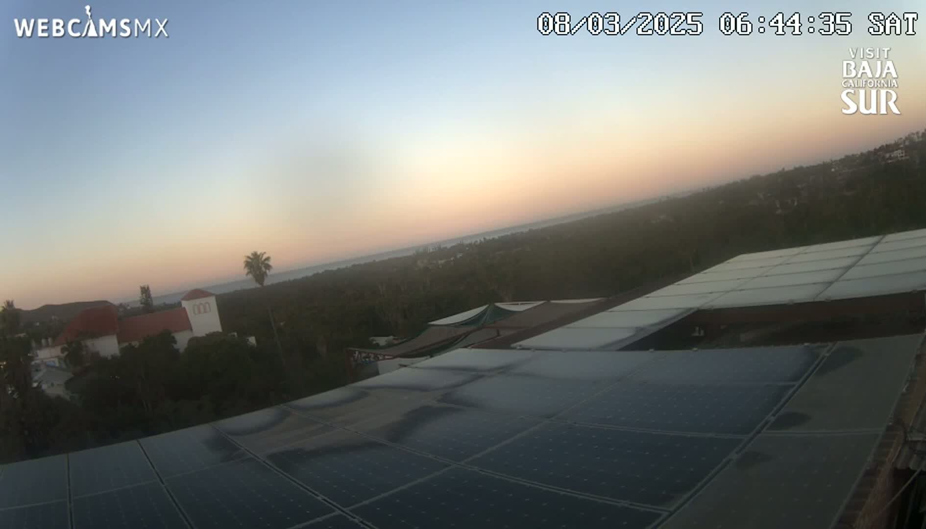 A sunrise illuminates a coastal landscape, viewed from a rooftop covered in solar panels, with buildings and vegetation visible in the foreground and a calm ocean in the distance.