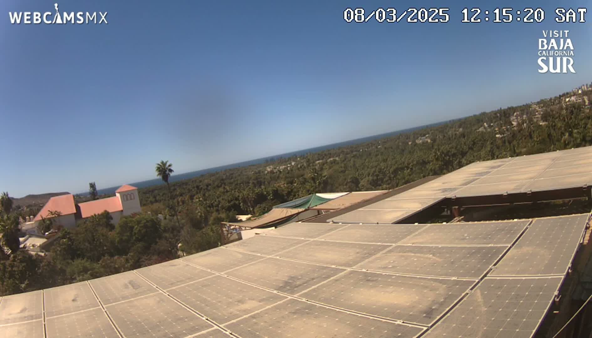 A large array of solar panels sits atop a building overlooking a distant ocean under a clear blue sky.