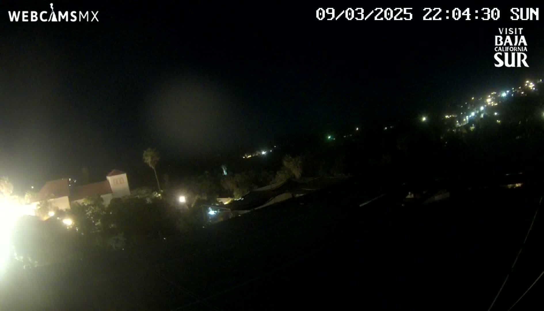 A nighttime view shows a town with scattered lights, a building with lights on, and a dark, clear sky.