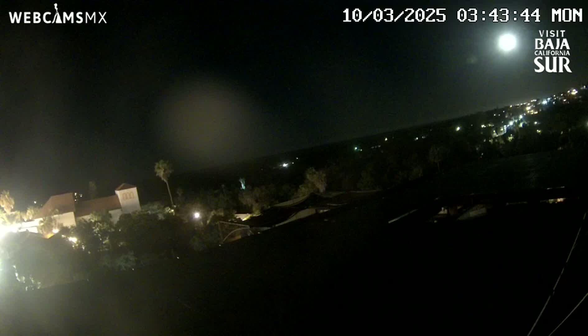 A nighttime, high-angle view shows a bright moon in the sky over a dark landscape with a few lights visible in the distance and a building illuminated closer to the camera.