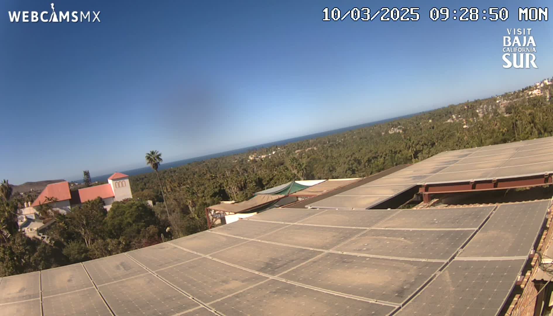 A large solar panel array sits atop a building overlooking a coastal town and ocean under a clear blue sky.