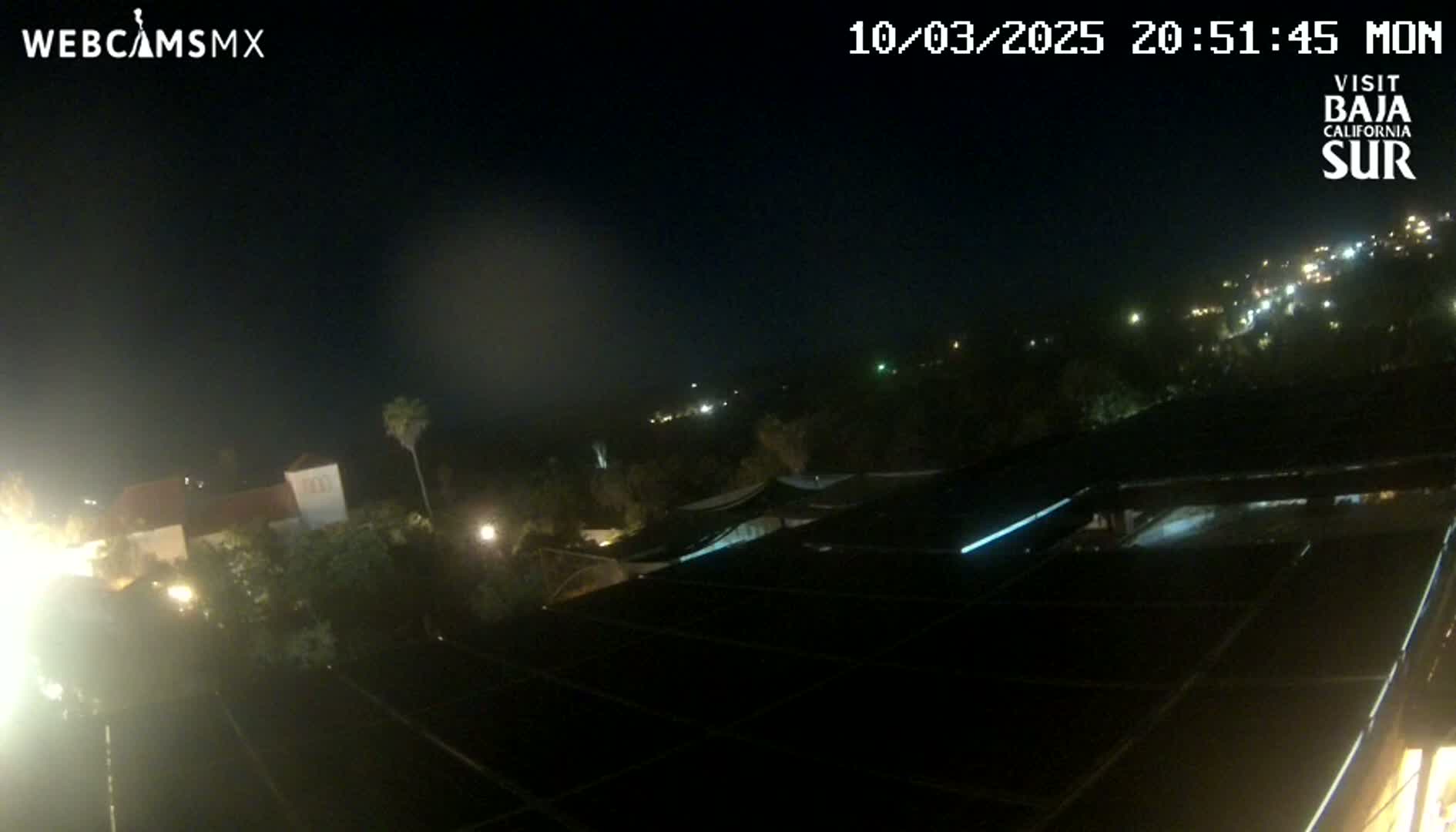 A nighttime view of a city with a dark sky, showing buildings and streetlights, as seen from a rooftop with solar panels.