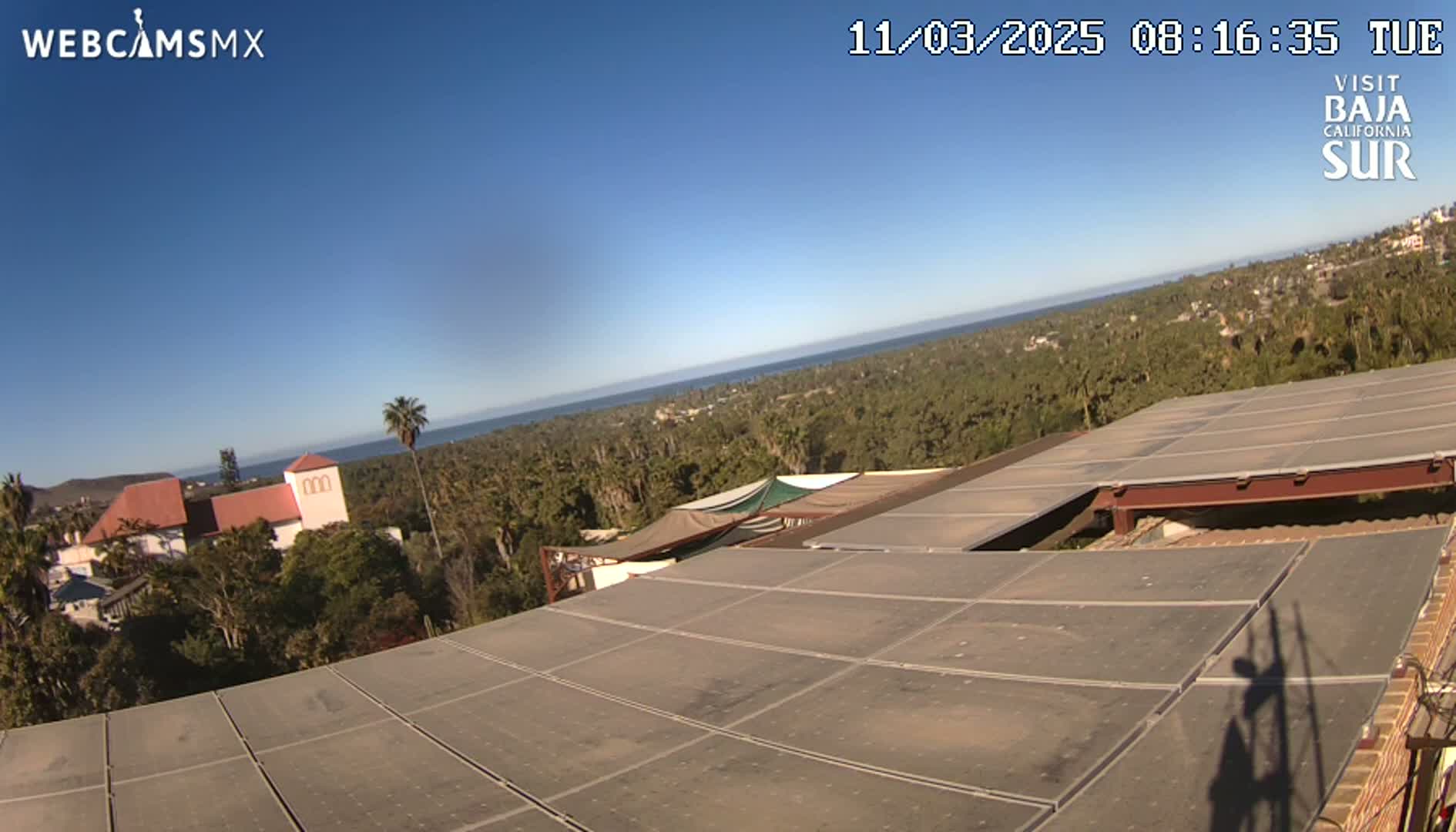 A large flat rooftop with solar panels overlooks a coastal town and ocean under a clear blue sky.