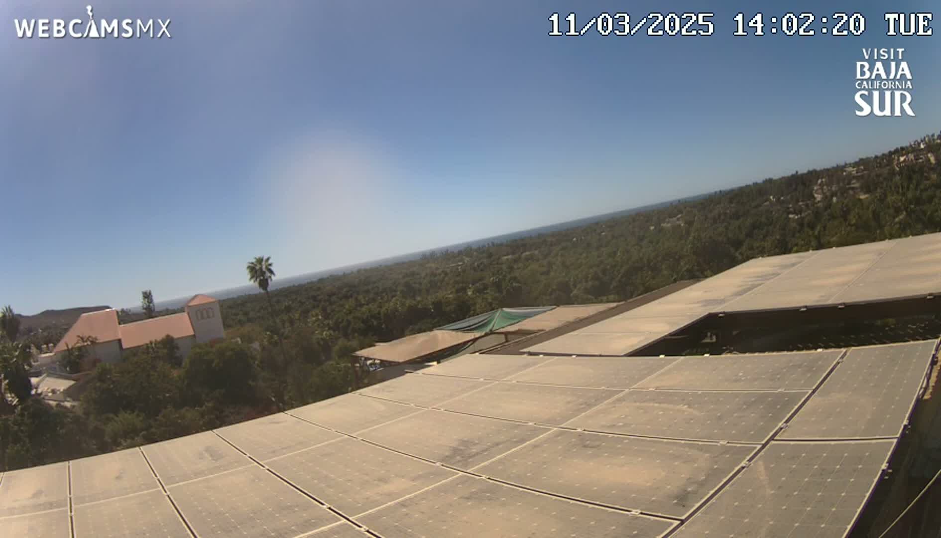 A large solar panel array sits atop a building overlooking a sunny, tree-covered landscape and a distant ocean.